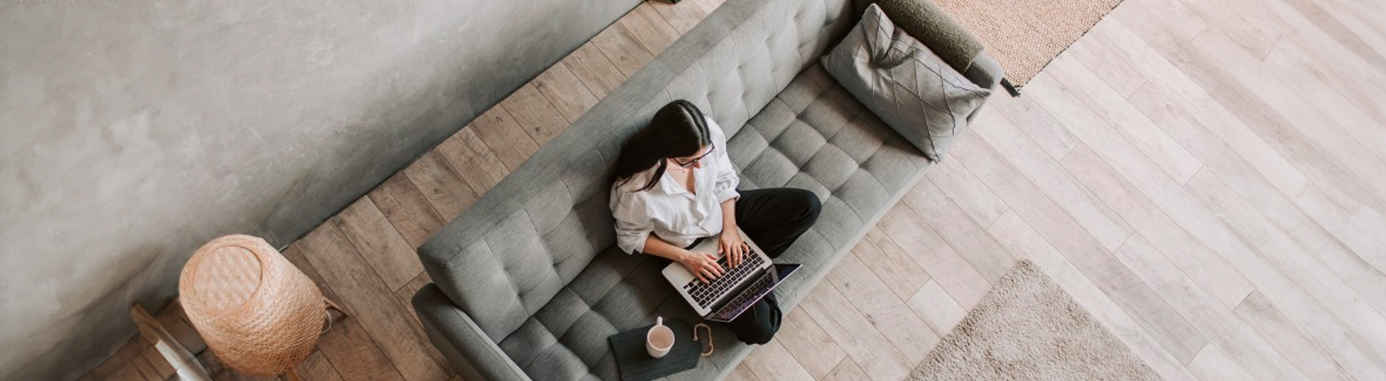 A woman sits on a sofa with her legs crossed working on a Mac with an empty cup of coffee.
